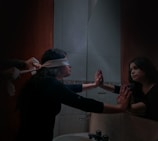 A person with a blindfold is standing in a dimly lit bathroom, facing a mirror with hands pressed against it. Another pair of hands is seen tying the blindfold around the person's head. The individual's reflection is visible in the mirror, creating a poignant scene.