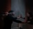 A person with a blindfold is standing in a dimly lit bathroom, facing a mirror with hands pressed against it. Another pair of hands is seen tying the blindfold around the person's head. The individual's reflection is visible in the mirror, creating a poignant scene.