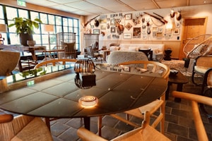 A cozy and eclectic interior features a mix of wooden and rattan furniture, including chairs and tables. A variety of framed pictures and mounted animal heads adorn the wall, creating a rustic and artistic atmosphere. Large windows allow natural light to fill the space, highlighting plants and decorative elements placed around the room.