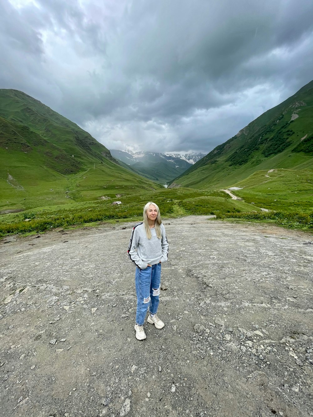 Mujer con chaqueta gris y jeans de mezclilla azul de pie en un camino de tierra gris durante el día