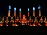 A nighttime scene featuring a monument with a central statue surrounded by large vertical pillars. These pillars are illuminated with blue lights at the top and display portraits of notable figures. Below the portraits, flowers and candles create a bright, warm glow. Silhouettes of people are gathered in the foreground, contributing to the solemn and respectful atmosphere.
