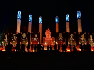 A nighttime scene featuring a monument with a central statue surrounded by large vertical pillars. These pillars are illuminated with blue lights at the top and display portraits of notable figures. Below the portraits, flowers and candles create a bright, warm glow. Silhouettes of people are gathered in the foreground, contributing to the solemn and respectful atmosphere.