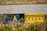 A weathered airport taxiway sign with a yellow background and black markings, displaying the letter 'A' and other symbols. The sign is surrounded by dry grass and low vegetation, with part of an airport runway visible in the background.