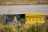 A weathered airport taxiway sign with a yellow background and black markings, displaying the letter 'A' and other symbols. The sign is surrounded by dry grass and low vegetation, with part of an airport runway visible in the background.