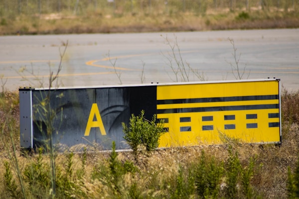 A weathered airport taxiway sign with a yellow background and black markings, displaying the letter 'A' and other symbols. The sign is surrounded by dry grass and low vegetation, with part of an airport runway visible in the background.