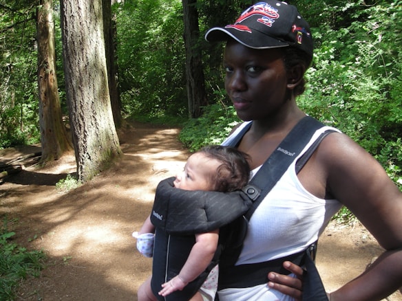A woman is carrying a baby in a carrier while walking through a lush, green forest path. She wears a cap and a white tank top. Tall trees and dense foliage surround the path, creating a serene and natural environment.