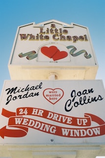 The image displays a sign for 'A Little White Chapel' featuring red hearts and ribbons. The lower section highlights that notable figures have married there, with names like Michael Jordan and Joan Collins. It also promotes a 24-hour drive-up wedding window.
