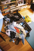 An open suitcase is filled with a variety of clothing items, including jeans, a red shirt, and a colorful garment. Surrounding the suitcase are household items like a yellow shopping bag and shelves filled with toys and books. The scene appears slightly cluttered, with some objects scattered on the floor.