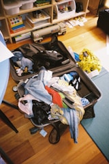 An open suitcase is filled with a variety of clothing items, including jeans, a red shirt, and a colorful garment. Surrounding the suitcase are household items like a yellow shopping bag and shelves filled with toys and books. The scene appears slightly cluttered, with some objects scattered on the floor.