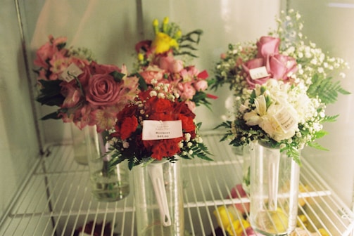 Bouquets of flowers are displayed in vases on shelves. The arrangements include roses, carnations, and baby's breath in various colors such as pink, red, and white. Price tags are visible on some of the bouquets. The setting appears to be inside a floral cooler, showcasing the bouquets for sale.