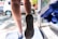 A close-up view of a person’s foot stepping onto a mat while a gloved hand holds a spray bottle, with liquid droplets visible near the nozzle. The background includes a blurred car and outdoor setting, suggesting a focus on cleaning or disinfection.