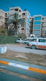 A multi-story residential or office building with a large number 30 displayed on one side. A palm tree is in the foreground along with other greenery. Two parked vehicles, including a white and red van showing signs of wear, are near the building. The scene has a clear blue sky and the sidewalk in front is marked with yellow and white stripes.
