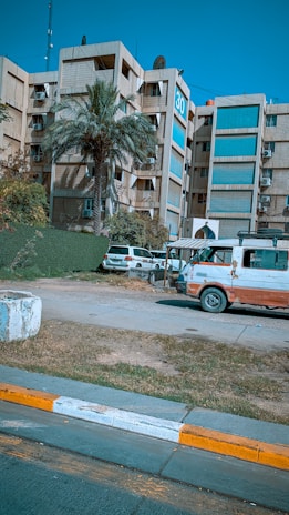 A multi-story residential or office building with a large number 30 displayed on one side. A palm tree is in the foreground along with other greenery. Two parked vehicles, including a white and red van showing signs of wear, are near the building. The scene has a clear blue sky and the sidewalk in front is marked with yellow and white stripes.