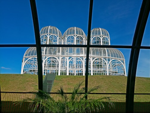 A large glass conservatory structure with multiple domes sits atop a grassy hill. The structure features arched windows and white frameworks. In the foreground, green plants can be seen inside another glass structure with visible black frames.