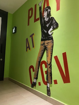 A mural features a stylized figure dressed in a black top and orange striped pants, posing with one hand on the hip and the other arm extended upward. The words 'PLAY AT C.G.V' are written in bold red and black letters on a bright green wall.