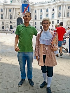 A man in a green polo shirt and jeans stands beside a person dressed in historical attire with a wig, glasses, and holding books. They are outside a grand architectural building with sculptures and people walking around.
