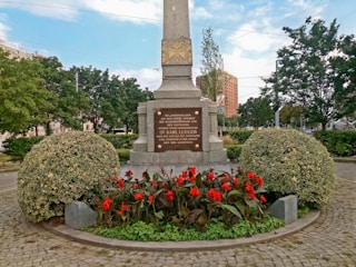 A monument surrounded by vibrant red flowers and neatly trimmed bushes stands in an outdoor setting. The stone base of the monument has inscriptions. Behind the monument, there are trees and a glimpse of buildings in the background under a partly cloudy sky.