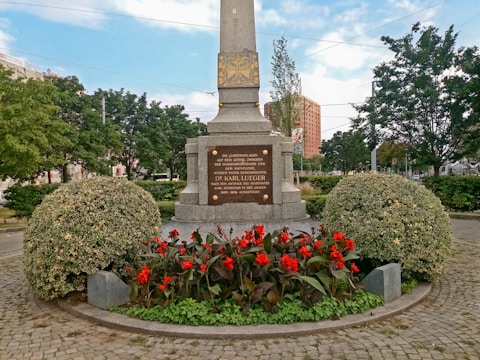 A monument surrounded by vibrant red flowers and neatly trimmed bushes stands in an outdoor setting. The stone base of the monument has inscriptions. Behind the monument, there are trees and a glimpse of buildings in the background under a partly cloudy sky.