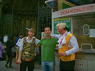 Three men are standing together in front of a ticket booth. Two of them are wearing costumes with traditional elements, featuring ornate vests and accessories, while the man in the middle is dressed casually in a green shirt and jeans. They appear to be smiling and interacting in a friendly manner, with one man having his arm around the central figure. Behind them is a decorative metal fence and signage related to a concert venue or historical site.