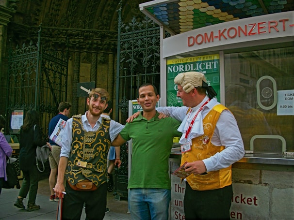 Three men are standing together in front of a ticket booth. Two of them are wearing costumes with traditional elements, featuring ornate vests and accessories, while the man in the middle is dressed casually in a green shirt and jeans. They appear to be smiling and interacting in a friendly manner, with one man having his arm around the central figure. Behind them is a decorative metal fence and signage related to a concert venue or historical site.