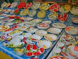 A display of various colorful ceramic plates and bowls adorned with floral and fruit patterns. The patterns include vibrant red flowers, yellow lemons, green leaves, and other nature-inspired designs. The arrangement is set on a blue tablecloth with a daisy pattern.