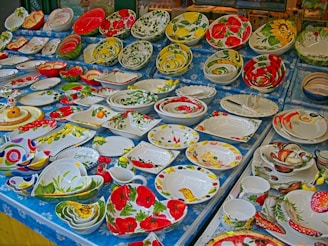 A display of various colorful ceramic plates and bowls adorned with floral and fruit patterns. The patterns include vibrant red flowers, yellow lemons, green leaves, and other nature-inspired designs. The arrangement is set on a blue tablecloth with a daisy pattern.