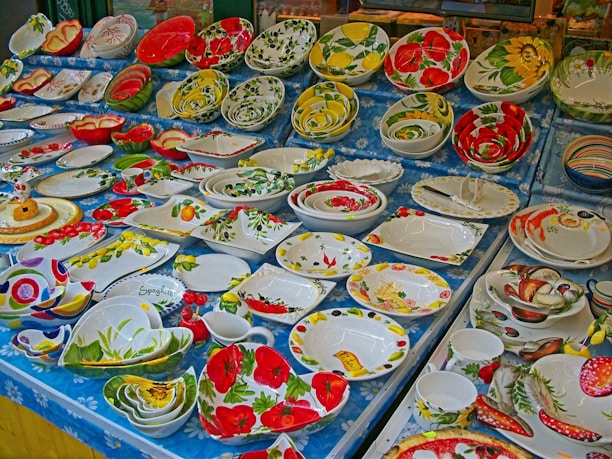 A display of various colorful ceramic plates and bowls adorned with floral and fruit patterns. The patterns include vibrant red flowers, yellow lemons, green leaves, and other nature-inspired designs. The arrangement is set on a blue tablecloth with a daisy pattern.