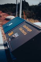 A tilted view of a dark blue garbage bin with the words 'Garbage Déchets' written in yellow. The number 36 is also visible. The setting appears to be outdoors, with natural surroundings including some blurred foliage and rocks.