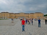 A large, historic palace with an impressive facade characterized by numerous windows and symmetrical architecture. The courtyard is filled with tourists walking and taking photos, with cobblestone paving under a cloudy sky.