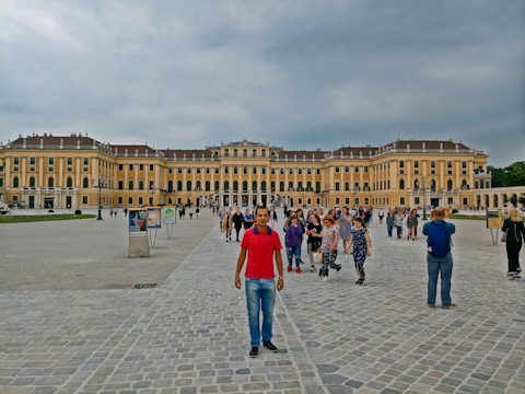 A large, historic palace with an impressive facade characterized by numerous windows and symmetrical architecture. The courtyard is filled with tourists walking and taking photos, with cobblestone paving under a cloudy sky.