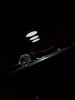 A person is playing pool in a dimly lit room, focusing intently on their shot. The pool table is illuminated by overhead lights, creating a dramatic effect. The person is wearing a plaid shirt, and the atmosphere is quiet and concentrated.