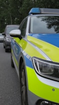 A police car with a blue and yellow color scheme is parked on the side of a road. The car is equipped with a roof-mounted light bar, and another vehicle is visible in the background. The scene takes place in a wooded area with trees lining the road.