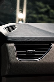 A close-up view of a car's interior focusing on the dashboard and air vent. The dashboard features a geometric pattern, and the air vent has a horizontal grille with a control knob. The background shows a partially blurred view with lights and shadows.