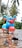 A colorful toy scooter is placed on a wet surface. The scooter is predominantly blue with red handlebars and front fender, featuring white wheels and yellow accents. Palm trees and lush greenery are visible in the blurry background.
