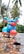A colorful toy scooter is placed on a wet surface. The scooter is predominantly blue with red handlebars and front fender, featuring white wheels and yellow accents. Palm trees and lush greenery are visible in the blurry background.
