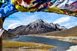 A serene landscape showcases a tranquil river flowing through a valley surrounded by towering, rugged mountains. The snow-capped peaks stand majestically under a partly cloudy sky. In the foreground, colorful prayer flags flutter in the breeze, adding a sense of spirituality and cultural significance to the scene.