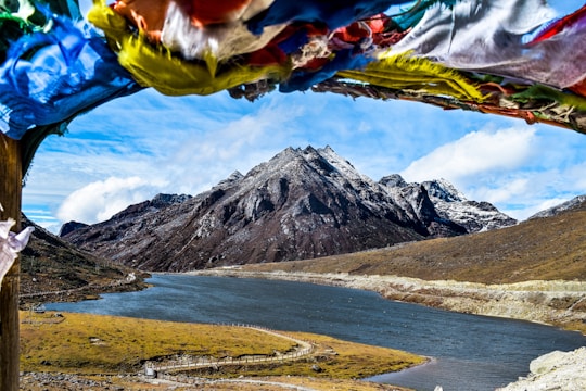 A serene landscape showcases a tranquil river flowing through a valley surrounded by towering, rugged mountains. The snow-capped peaks stand majestically under a partly cloudy sky. In the foreground, colorful prayer flags flutter in the breeze, adding a sense of spirituality and cultural significance to the scene.