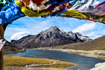 A serene landscape showcases a tranquil river flowing through a valley surrounded by towering, rugged mountains. The snow-capped peaks stand majestically under a partly cloudy sky. In the foreground, colorful prayer flags flutter in the breeze, adding a sense of spirituality and cultural significance to the scene.