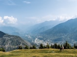A vast mountainous landscape features a valley below with a winding river. Pine trees are scattered along the foreground, and two people are walking on the grass. Colorful prayer flags are strung across the scene, while fluffy clouds hover in the sky above the distant peaks.