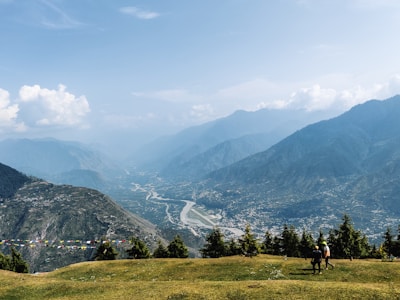 A vast mountainous landscape features a valley below with a winding river. Pine trees are scattered along the foreground, and two people are walking on the grass. Colorful prayer flags are strung across the scene, while fluffy clouds hover in the sky above the distant peaks.