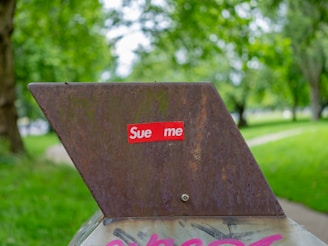 A rusty metal object with a prominent sticker displaying 'Sue me' in white text on a red background. The object is set against a backdrop of a lush green park with trees and a walkway.