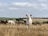 A group of goats and sheep grazing in a harvested field under a partly cloudy sky. The central goat is prominently facing towards the camera, while other animals feed on the dry grass. The backdrop features sprawling open fields with low-lying hills and a few scattered trees.