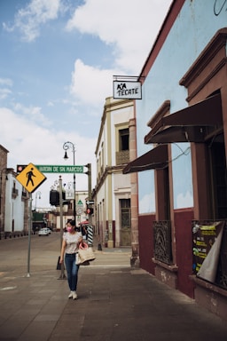 A woman wearing a mask walks down a street lined with historic buildings. The sky is partly cloudy, and multiple street signs are visible, including one for 'Jardin de San Marcos.' A 'Tecate' beer sign hangs above the sidewalk.