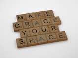 Wooden letter tiles are arranged on a flat surface to spell out the phrase 'MAKE GRACE YOUR SPACE'. Each tile has a letter with a corresponding point value at the bottom right corner, typical of a Scrabble game.