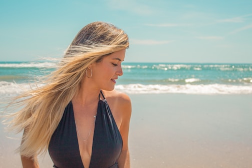 A woman with long blonde hair stands on a sandy beach with the ocean in the background. She is looking to her side, wearing a black swimsuit. The sunlight illuminates her hair, creating a warm and serene atmosphere.
