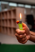 A hand is holding a lit lighter with a visible flame. The lighter is a bright green color and is being held against a blurred background that includes what appears to be shelving or storage units.