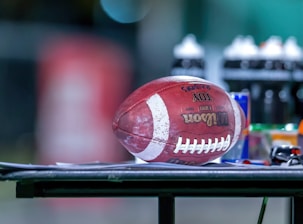 A football is resting on a flat surface, surrounded by various sports bottles and gear. The background is blurred, emphasizing the football and the objects on the table.