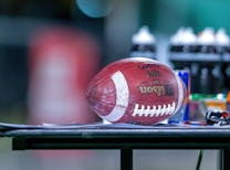 A football is resting on a flat surface, surrounded by various sports bottles and gear. The background is blurred, emphasizing the football and the objects on the table.
