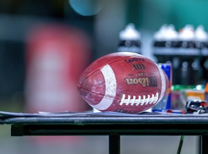 A football is resting on a flat surface, surrounded by various sports bottles and gear. The background is blurred, emphasizing the football and the objects on the table.