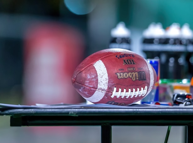 A football is resting on a flat surface, surrounded by various sports bottles and gear. The background is blurred, emphasizing the football and the objects on the table.